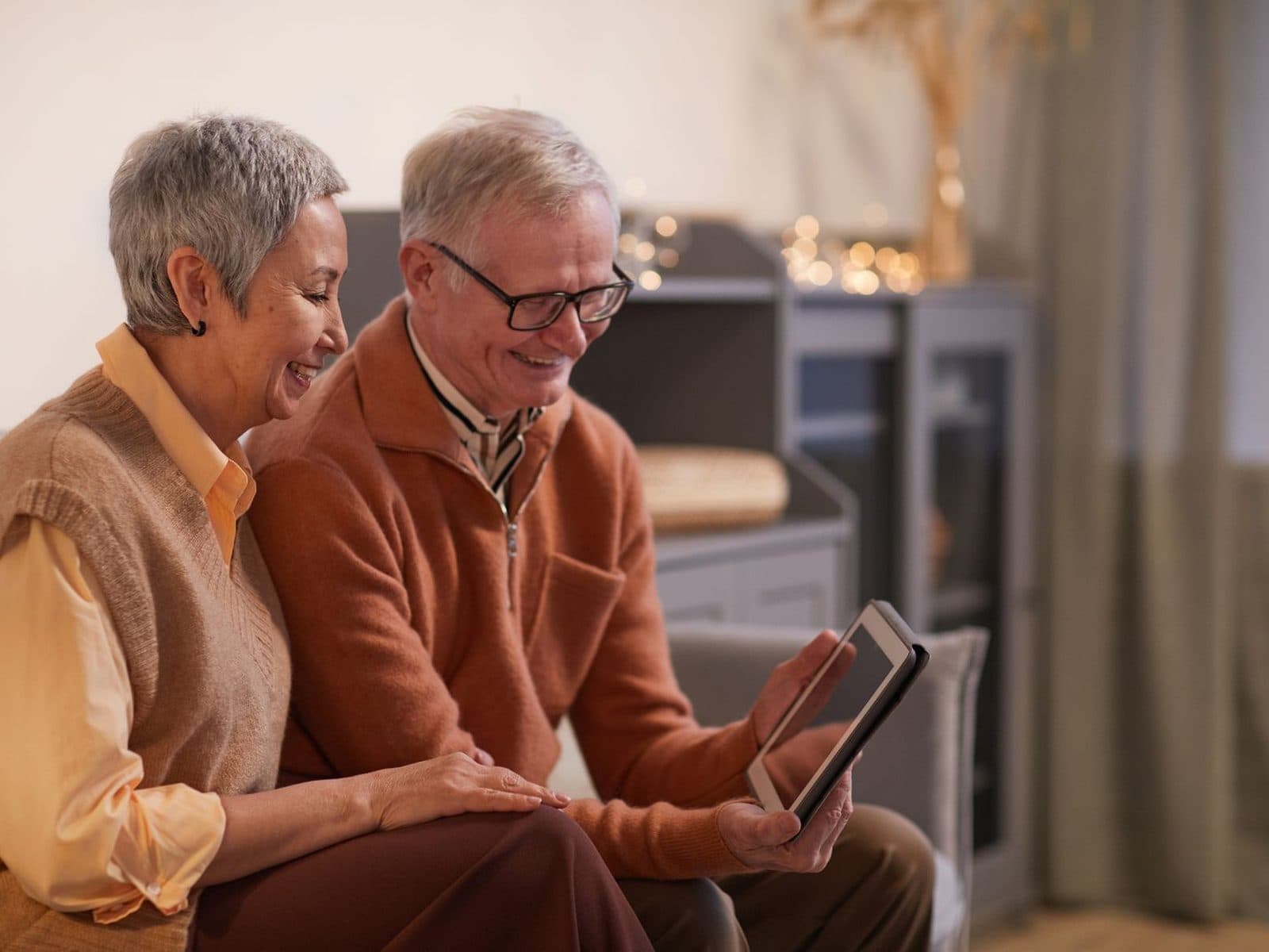An older couple side by side in their front room, smiling at the tablet they are booking on